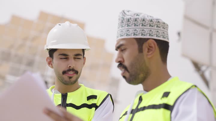 Two Arab Gulf engineers from Oman supervise solar power stations, carrying a plan and engineering design, wearing helmets and safety vests, overseeing the work, satellite, solar power systems, exchanging experience and opinions among colleagues.