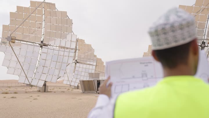 تصوير من الخلف لمهندس عربي خليجي عماني يرتدي الدشداشة والكمة و سترة الحماية ينظر بإيماءات السرور يحمل بيده مخططات وتصاميم هندسية,  مهن ووظائف للشباب في سلطنة عمان