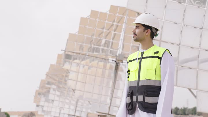 النظر الى شىء ما, الإشراف على محطات توليد الكهرباء, استخدام التقنية والتكنولوجيا في المشاريع المستقبلية, مهندس طاقة عربي خليجي عمانى يقف بأيدى متقاطعة، إرتداء خوذة وسترة الحماية الخاصة بالعمل