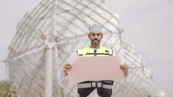 الاهتمام بشؤون العمل, تقدم و تطور سلطنة عمان على يد أبناء الوطن, مهندس عربي خليجي عماني يرتدي الدشداشة والكمة و سترة الحماية ينظر بإيماءات السرور  يحمل بيده مخططات وتصاميم هندسية, التدقيق والإشراف على العمل