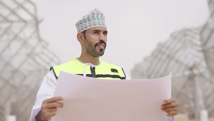 مهندس عربي خليجي عماني يرتدي الدشداشة والكمة  و سترة الحماية ينظر بإيماءات السرور  يحمل بيده مخططات وتصاميم هندسية, التدقيق والإشراف على العمل,  مفهوم هندسة الطاقة