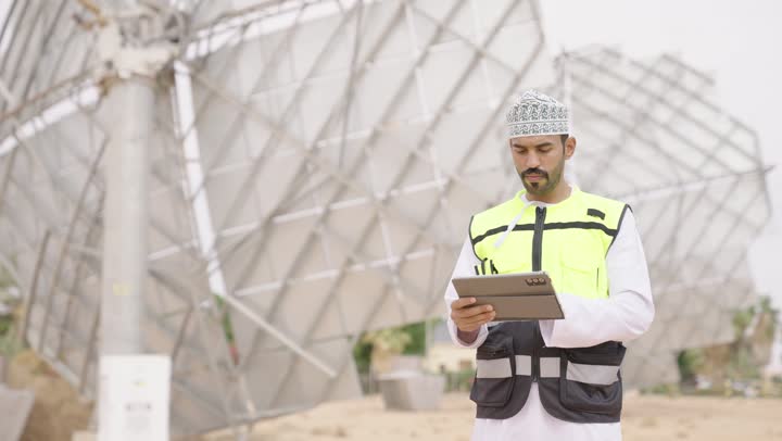 The concept of energy engineering, an Arab Gulf Omani engineer wearing a dishdasha and kumma and a safety vest looks with gestures of joy holding a tablet in his hand, wearing a helmet and safety vest, auditing and supervising the work.