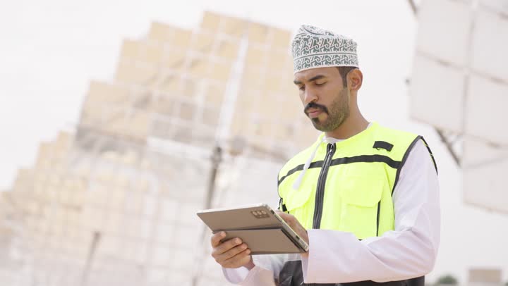 استخدام جهاز تقني حديث متطور في مجال الهندسة, مهندس عربي خليجي عماني يرتدي الدشداشة والكمة و سترة الحماية يحمل بيده التابلت، إرتداء خوذة وسترة الحماية الخاصة بالعمل، التدقيق والإشراف على العمل، الطاقة المتجددة