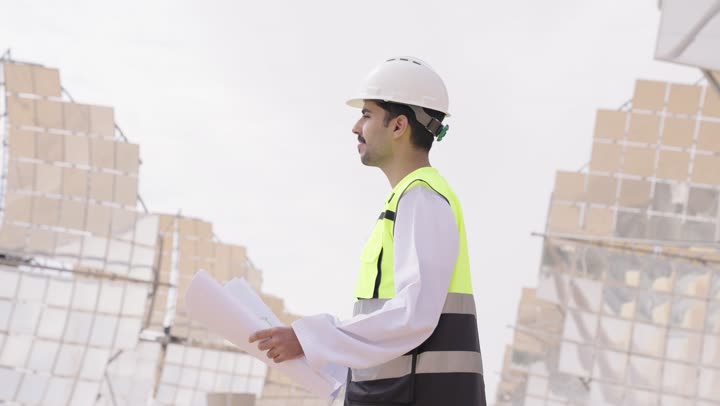 مفهوم هندسة الطاقة، مهن ووظائف عمانية، محطات الطاقة الشمسية, التدقيق والإشراف على العمل, تصوير من الجانب لمهندس عربي خليجي عمانى يرتدي سترة و خوذة الحماية يقف في مقر العمل و يحمل مخطط هندسى