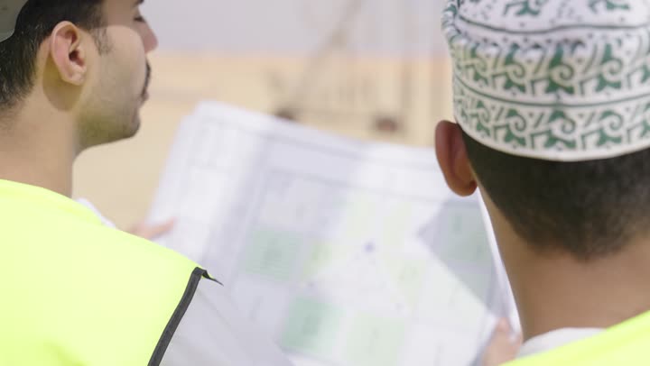 تصوير من الخلف لمهندسان عربيان خليجيان عمانيان يشرفان على محطات الطاقة الشمسية ، إرتداء خوذة وسترة الحماية ، الإشراف على العمل، ستلايت، أنظمة الطاقة الشمسية,  تبادل الخبرة و الرأى,