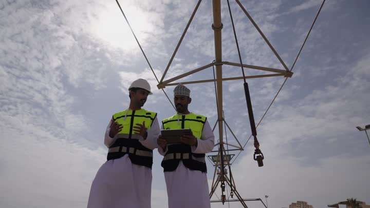 حوار ونقاش عمل ، محطات الطاقة الشمسية المتجددة، الواح وستلايت انتاج الطاقة الكهربائية, صورة من الاسفل لمهندسان عربيان خليجيان عمانيان يشرفان على محطات الطاقة الشمسية في عمان، إرتداء خوذة وسترة الحماية الخاصة بالعمل