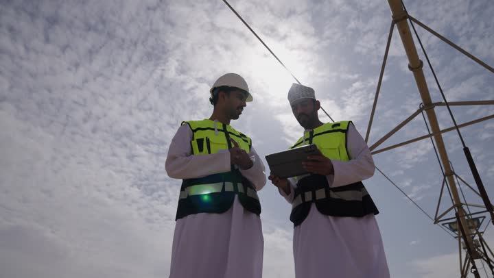صورة من الاسفل لمهندسان عربيان خليجيان عمانيان يشرفان على محطات الطاقة الشمسية في السعودية، إرتداء خوذة وسترة الحماية الخاصة بالعمل، مخططات وتصاميم هندسية، حوارات ونقاشات عمل ، محطات الطاقة الشمسية المتجددة، الواح وستلايت انتاج الطاقة الكهربائية