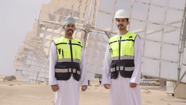 محطة الطاقة الشمسية المتجددة، الواح وستلايت انتاج الطاقة الكهربائية, مهندسان عربيان خليجيان عمانيان يشرفان على محطات الطاقة الشمسية يقفان بأيدى متقاطعة, إرتداء خوذة وسترة الحماية الخاصة بالعمل، مخططات وتصاميم هندسية، حوارات ونقاشات عمل