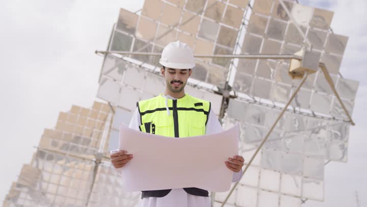 Auditing and supervising work, an Arab Gulf Omani engineer wearing a vest and safety helmet standing at the workplace holding an engineering plan, the concept of energy engineering, Omani professions and jobs, solar power stations.