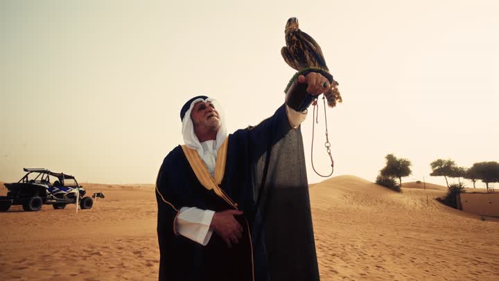 الاهتمام بتربية وتدريب الطيور الجارحة, رجل عربي خليجي إماراتي مسن يرتدي الكندورة البيضاء والبشت الأسود والشماغ مع العقال يقف فى الصحراء يحمل على يدية صقر، ايماءات السعادة و السرور