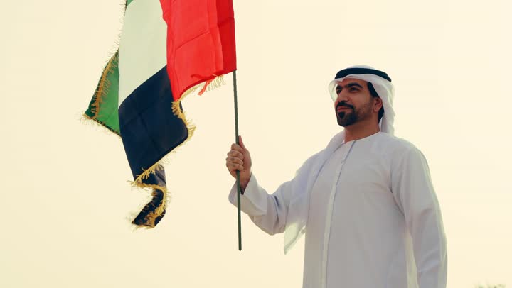 حب الوطن والاعتزاز به، اليوم الوطني الاماراتي 1971، شاب عربي خليجي إماراتي يرتدي الكندورة والغترة يمسك بيده علم الإمارات العربية المتحدة يقف الصحراء, رفع راية الوطن في يوم العلم 3 نوفمبر