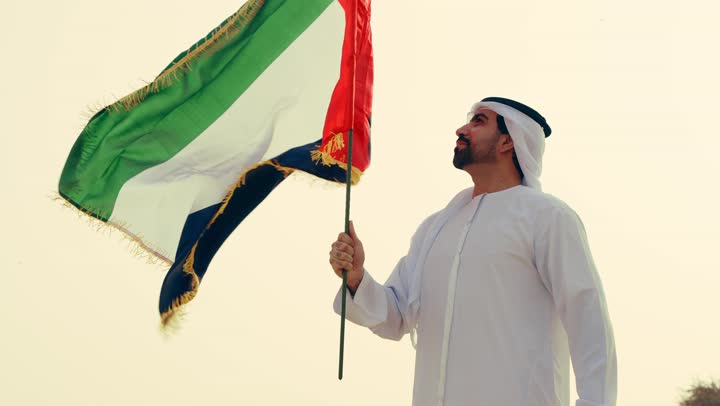 Celebrating the anniversary of the union's establishment on December 2, love for the homeland and pride in it, the UAE National Day 1971, a young Arab Gulf Emirati wearing the kandura and ghutrah holding the flag of the United Arab Emirates standing in the desert.