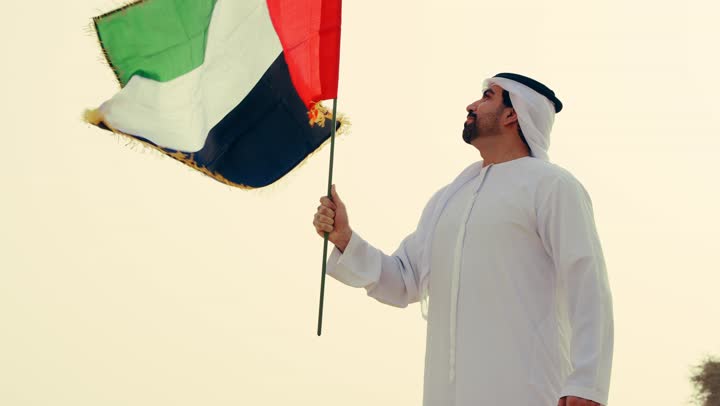 النظر الى راية الوطن في يوم العلم 3 نوفمبر بفخر واعتزاز، استذكار اليوم الوطني الاماراتي 1971، تأسيس الاتحاد 2 ديسمبر، شاب عربي خليجي إماراتي يرتدي الكندورة والغترة يمسك بيده علم الإمارات العربية المتحدة يقف الصحراء