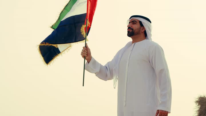 شاب عربي خليجي إماراتي يرتدي الكندورة والغترة يمسك بيده علم الإمارات العربية المتحدة و يرفع اصبع يديه بفخر، حب الوطن والاعتزاز به,  استذكار اليوم الوطني الاماراتي 1971، تأسيس الاتحاد 2 ديسمبر