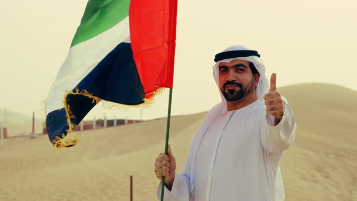 Commemoration of the UAE National Day 1971, the establishment of the union on December 2, a young Arab Gulf Emirati wearing the kandura and ghutrah holding the flag of the United Arab Emirates in his hand and raising his finger proudly, love for the homeland and pride in it.