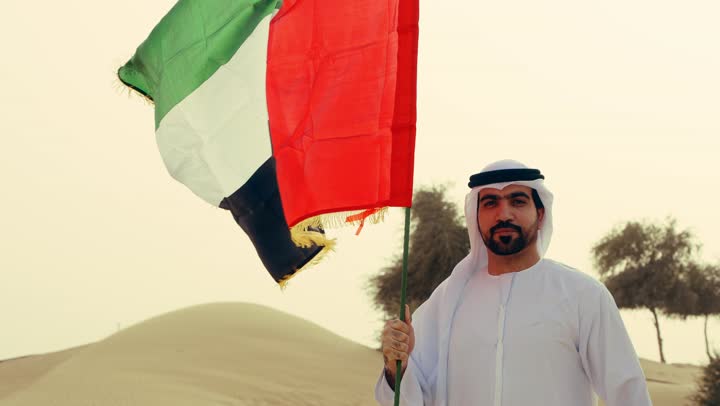 رفع راية الوطن في يوم العلم 3 نوفمبر، استذكار اليوم الوطني الاماراتي 1971، تأسيس الاتحاد 2 ديسمبر، شاب عربي خليجي إماراتي يرتدي الكندورة والغترة يمسك بيده علم الإمارات العربية المتحدة يقف الصحراء في وقت النهار، حب الوطن والاعتزاز به، خلفية الطبيعة الصحراوية