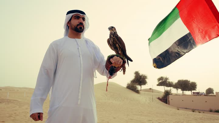 تنمية قدرات الطيور من خلال تربيتها, مكانة الصقور في التاريخ الاماراتي, رجل عربي خليجي اماراتي يرتدي الكندورة و الغتره البيضاء يقف فى الصحراء يحمل على يده طائر الصقرينظر الى الكاميرا بتعابير السرور، تدريب الصقر الجارح