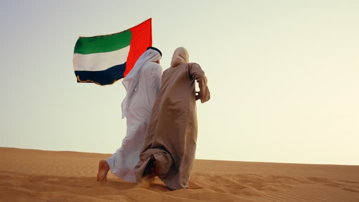 تصوير من الخلف لرجل عربي خليجي اماراتي يرتدي الكندورة والغترة يتجول فى الصحراء برفقة زوجته يمسك بيدية علم الامارات، ايماءات السعادة والفخر والاعتزاز، اليوم الوطني الإماراتي 2 ديسمبر