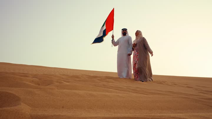 ذكرى استقلال دولة الإمارات العربية المتحدة, العيد الوطني الإماراتي، رجل عربي خليجي اماراتي يرتدي الكندورة والغترة يتجول فى الصحراء برفقة زوجته يمسك بيدية علم الامارات، ايماءات السعادة والفخر والاعتزاز، اليوم الوطني الإماراتي 2 ديسمبر