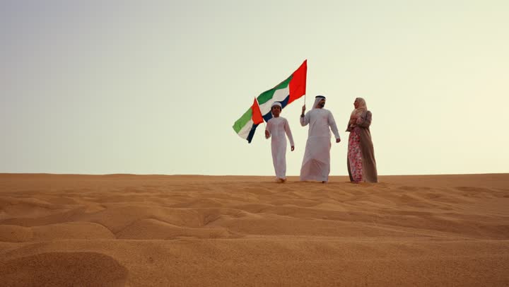 عائلة عربية خليجية إماراتية ترتدي الزي التقليدي تمشى في الصحراء وترفع علم دولة الإمارات العربية المتحدة، أجواء تراثية تعكس الأصالة والانتماء الوطني, مفهوم الفخر والوحدة الوطنية، الاحتفال بالهوية الوطنية والانتماء