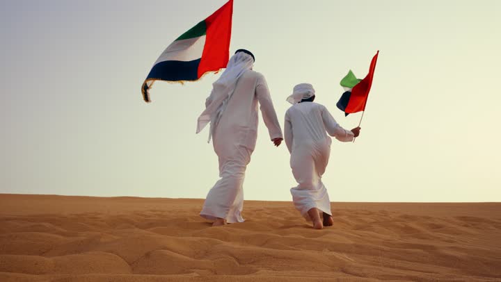 نصوير من الخلف لرجل عربي خليجي اماراتي يرتدي الكندورة والغترة يقف فى الصحراء برفقة ابنه يمسك بيدية علم الامارات, مفهوم الفخر والوحدة الوطنية،أجواء تراثية تعكس الأصالة والانتماء الوطني