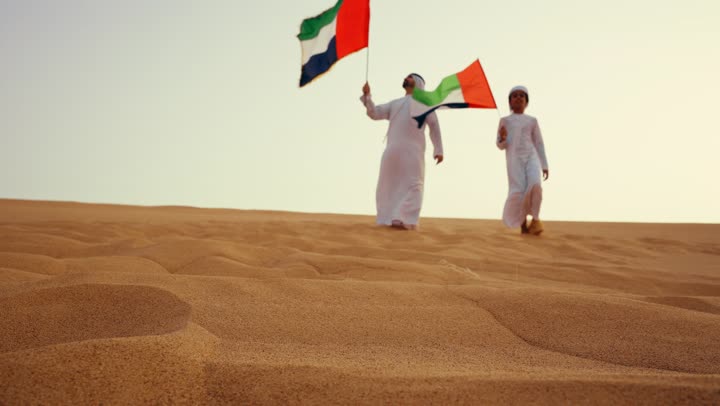 The concept of pride and national unity, a heritage atmosphere that reflects authenticity and national belonging, an Arab Gulf Emirati man wearing the kandura and ghutrah stands in the desert with his son holding the UAE flag in his hands.