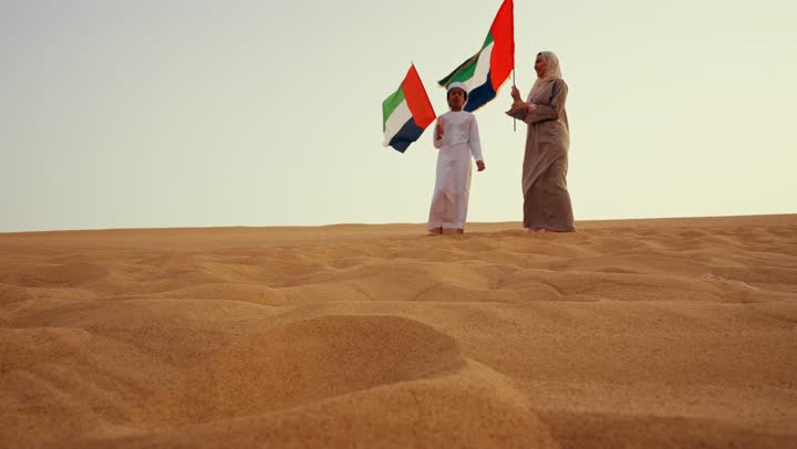 أجواء تراثية تعكس الأصالة والانتماء الوطني, مفهوم الفخر والوحدة الوطنية، الاحتفال بالهوية الوطنية والانتماء, امراة عربية خليجية اماراتية ترتدي العباءة والحجاب ترفع علم الإمارات في الصحراء مع ولدها فى الصحراء