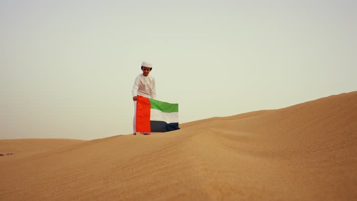 الاحتفال بالهوية الوطنية والانتماء، طفل عربي خليجي إماراتي يرتدي الكندورة و الغترة البيضاء يمسك بيده علم الإمارات العربية المتحدة يركض في الصحراء, حب الوطن والاعتزاز به, رفع راية الوطن في يوم العلم 3 نوفمبر، استذكار اليوم الوطني الاماراتي 1971