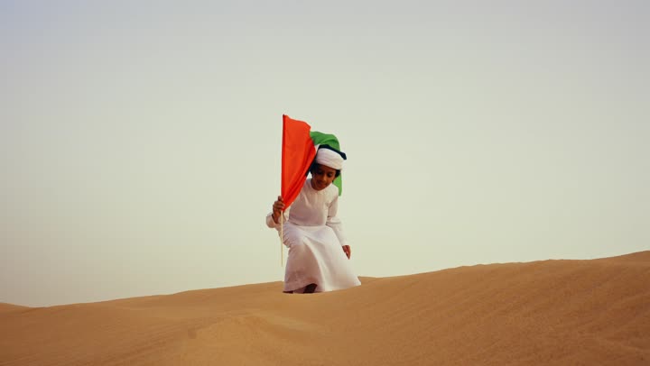 Gestures of happiness, pride, and honor, an Arab Gulf Emirati child wearing a kandura and a white ghutrah holds the flag of the United Arab Emirates in his hand while running in the desert, love for the homeland and pride in it, raising the flag of the nation on Flag Day, November 3, commemorating the UAE National Day 1971.