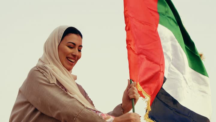 The concept of love for the homeland and belonging to it, celebrating the UAE National Day on December 2, commemorating the independence of the United Arab Emirates, an Arab Gulf Emirati woman wearing an abaya and hijab raising the UAE flag during the day, wandering in the desert with gestures of happiness, pride, and honor.