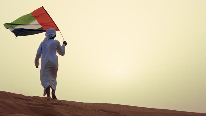 الاحتفال بالهوية الوطنية والانتماء, تصوير من الخلف لطفل عربي خليجي إماراتي يرتدي الكندورة و الغترة البيضاء يمسك بيده علم الإمارات العربية المتحدة يركض في الصحراء, رفع راية الوطن في يوم العلم 3 نوفمبر، استذكار اليوم الوطني الاماراتي 1971