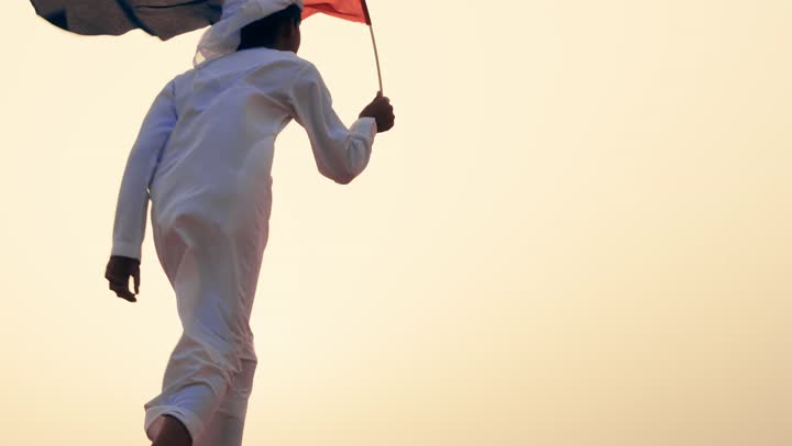 تصوبر من الخلف لطفل عربي خليجي إماراتي يرتدي الكندورة و الغترة البيضاء يمسك بيده علم الإمارات العربية المتحدة يركض في الصحراء, حب الوطن والاعتزاز به, رفع راية الوطن في يوم العلم 3 نوفمبر، استذكار اليوم الوطني الاماراتي 1971