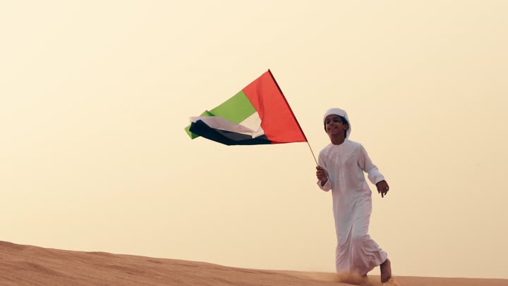 ذكرى استقلال دولة الإمارات العربية المتحدة, طفل عربي خليجي إماراتي يرتدي الكندورة و الغترة البيضاء يمسك بيده علم الإمارات العربية المتحدة يركض في الصحراء, حب الوطن والاعتزاز به, رفع راية الوطن في يوم العلم 3 نوفمبر، استذكار اليوم الوطني الاماراتي 1971