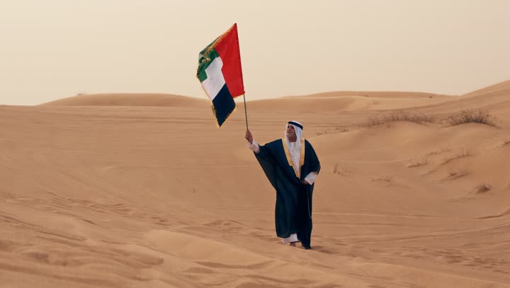 Wearing traditional folk costumes to celebrate the anniversary of the establishment of the union on December 2, an Arab Gulf Emirati man wearing a kandura and bisht with a white ghutrah holds the flag of the United Arab Emirates in his hand while standing in the desert, commemorating the UAE National Day 1971.