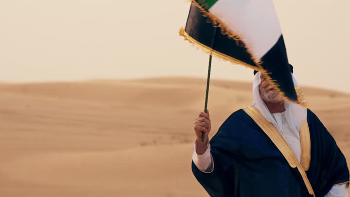An Arab Gulf Emirati man wearing a kandura and bisht with a white ghutrah is holding the flag of the United Arab Emirates while standing in an archaeological area in the desert. His love and pride for the homeland are evident as he raises the national flag on Flag Day, November 3, in commemoration of the UAE National Day 1971.