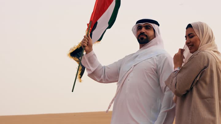 Gestures of happiness, pride, and honor, the UAE National Day on December 2, the anniversary of the independence of the United Arab Emirates, the UAE National Holiday, an Arab Gulf Emirati man wearing the kandura and ghutrah stands in the desert with his wife holding the UAE flag in his hands.