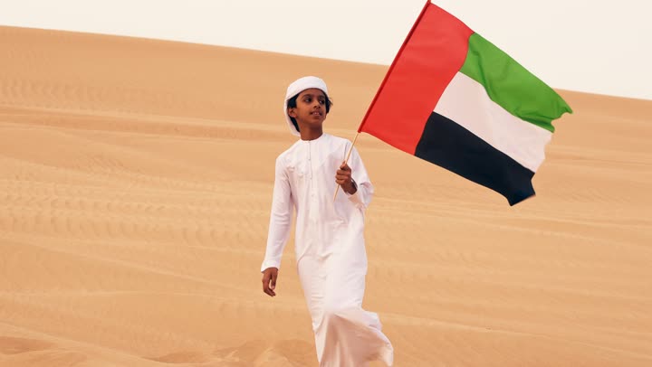 An Emirati Gulf Arab child wearing a kandura and a white ghutrah is holding the flag of the United Arab Emirates while standing in the desert, expressing love and pride for the homeland, commemorating the founding of the federation on December 2, raising the national flag on Flag Day, November 3, and remembering the UAE National Day in 1971.
