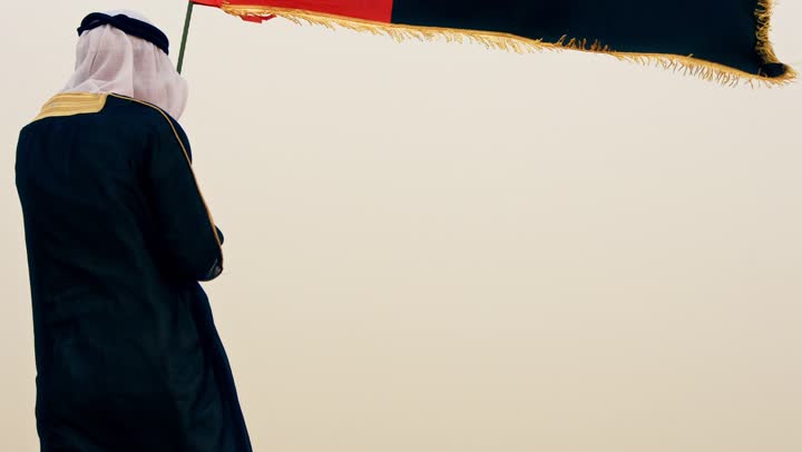 تصوير من الخلف لرجل عربي خليجي إماراتي يرتدي الكندورة والبشت مع الغترة البيضاء يمسك بيده علم الإمارات العربية المتحدة يقف في الصحراء,  تأسيس الاتحاد 2 ديسمبر, رفع راية الوطن في يوم العلم 3 نوفمبر، استذكار اليوم الوطني الاماراتي 1971،