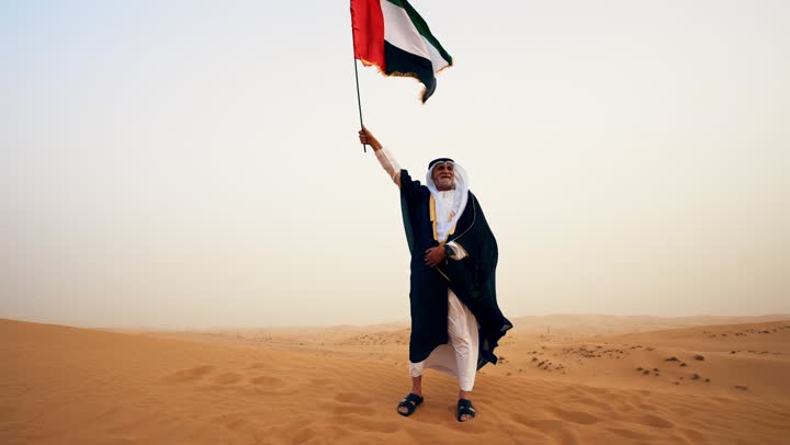 استذكار اليوم الوطني الاماراتي 1971، رجل عربي خليجي إماراتي يرتدي الكندورة والبشت مع الغترة البيضاء يمسك بيده علم الإمارات العربية المتحدة يقف في الصحراء,  تأسيس الاتحاد 2 ديسمبر, رفع راية الوطن في يوم العلم 3 نوفمبر،