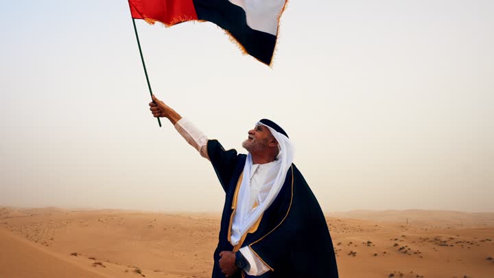 Establishment of the Union on December 2, raising the flag of the nation on Flag Day, November 3, commemorating the UAE National Day 1971, an Arab Gulf Emirati man wearing the kandura and bisht with a white ghutrah holding the flag of the United Arab Emirates standing in the desert.