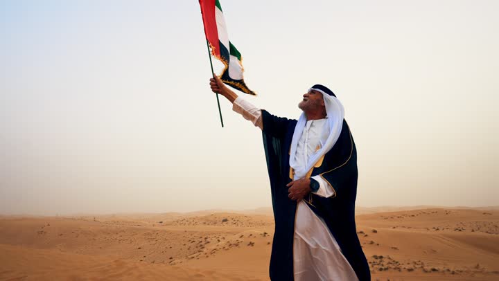 Raising the flag of the homeland on Flag Day, November 3, commemorating the UAE National Day 1971, the establishment of the union on December 2. An Arab Gulf Emirati man wearing the kandura and bisht with a white ghutrah holds the flag of the United Arab Emirates in his hand while standing in the desert.