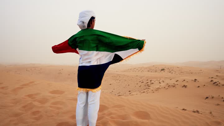 A rear view of an Emirati Gulf Arab child wearing a kandura and a white ghutrah, holding the flag of the United Arab Emirates in his hand, standing in the desert, love for the homeland and pride in it, the anniversary of the founding of the union on December 2, raising the flag of the homeland on Flag Day, November 3.