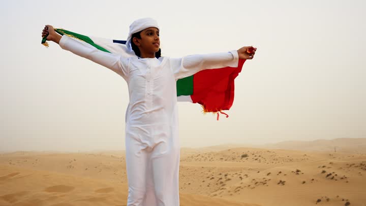 ذكرى تأسيس الاتحاد 2 ديسمبر، رفع راية الوطن في يوم العلم 3 نوفمبر، استذكار اليوم الوطني الاماراتي 1971، طفل عربي خليجي إماراتي يرتدي الكندورة و الغترة البيضاء يمسك بيده علم الإمارات العربية المتحدة يقف في الصحراء, حب الوطن والاعتزاز به