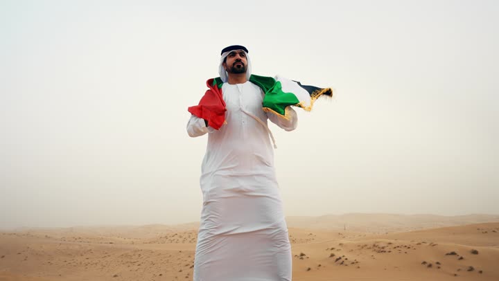 رجل عربي خليجي إماراتي يرتدي الكندورة و الغترة البيضاء يمسك بيده علم الإمارات العربية المتحدة يقف في الصحراء, حب الوطن والاعتزاز به، ذكرى تأسيس الاتحاد 2 ديسمبر، رفع راية الوطن في يوم العلم 3 نوفمبر، استذكار اليوم الوطني الاماراتي 1971،