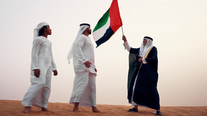 ارتداء الازياء الشعبية التقليدية للاحتفال بذكرى تأسيس الاتحاد 2 ديسمبر، يوم العلم 3 نوفمبر, حب الوطن والاعتزاز به, رجلان عربيان خليجيان اماراتيان يرتديان الزي التقليدي يرفعان علم الامارات العربية المتحدة في الصحراء