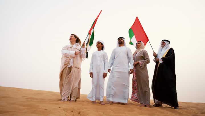 الاحتفال بالهوية الوطنية والانتماء, عائلة عربية خليجية إماراتية ترتدي الزي التقليدي تقف في الصحراء وترفع علم دولة الإمارات العربية المتحدة، أجواء تراثية تعكس الأصالة والانتماء الوطني, مفهوم الفخر والوحدة الوطنية، الاحتفال بالهوية الوطنية والانتماء