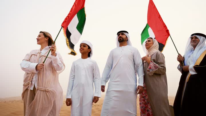The concept of love for the homeland and belonging to it, an Arab Gulf Emirati family dressed in traditional attire standing in the desert and raising the flag of the United Arab Emirates, a heritage atmosphere reflecting authenticity and national belonging, the concept of pride and national unity, celebrating national identity and belonging.