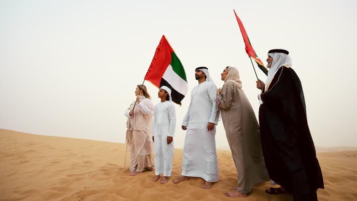 النظر الى شيء ما, عائلة عربية خليجية إماراتية ترتدي الزي التقليدي تقف في الصحراء وترفع علم دولة الإمارات العربية المتحدة، أجواء تراثية تعكس الأصالة والانتماء الوطني, مفهوم الفخر والوحدة الوطنية، الاحتفال بالهوية الوطنية والانتماء