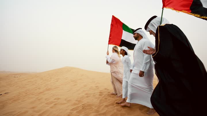 عائلة عربية خليجية إماراتية ترتدي الزي التقليدي تقف في الصحراء وترفع علم دولة الإمارات العربية المتحدة، أجواء تراثية تعكس الأصالة والانتماء الوطني, مفهوم الفخر والوحدة الوطنية، الاحتفال بالهوية الوطنية والانتماء,