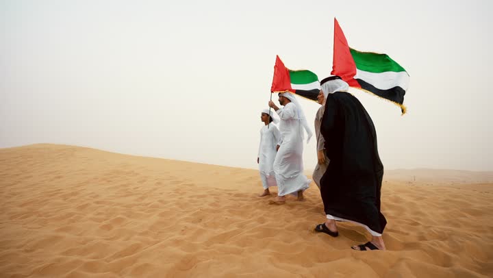 The concept of pride and national unity, celebrating national identity and belonging, an Emirati Gulf Arab family dressed in traditional attire standing in the desert and raising the flag of the United Arab Emirates, a heritage atmosphere reflecting authenticity and national belonging.